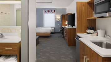 Una habitación de hotel moderna con kitchenette, una cama, un televisor y un baño con espejo y toallero.
