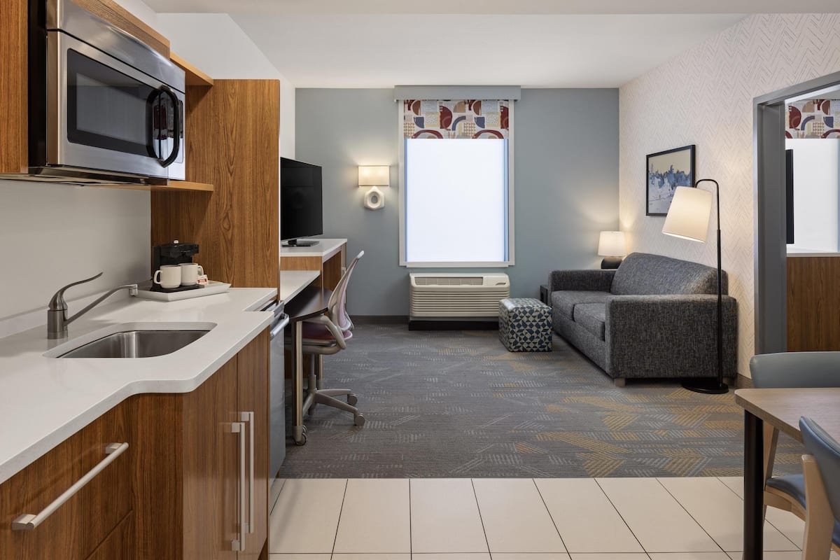 A compact hotel room with a kitchenette, a gray sofa, a small table, a TV, and a window with patterned curtains.