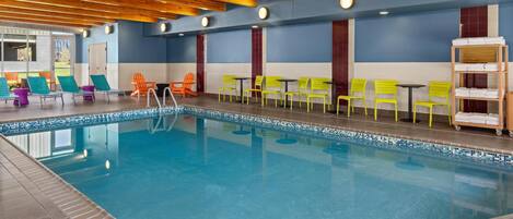 Una piscina interior con borde de azulejos, vigas de techo de madera y zonas de estar con sillas y mesas.