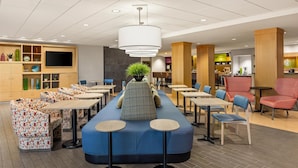 A modern lounge area with seating arrangements, a central plant, and a flat-screen TV mounted on the wall. - Home2 Suites by Hilton Roseville Minneapolis (Roseville)
