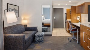 A hotel room with a sofa, a small ottoman, a desk, and a kitchenette. - Home2 Suites by Hilton Roseville Minneapolis (Roseville)