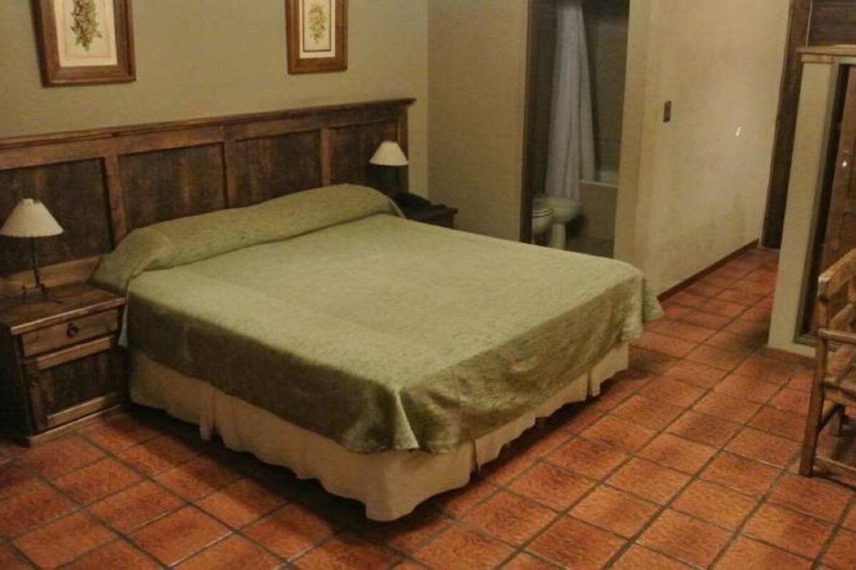 Una camera da letto con testiera in legno, un letto singolo, un comodino con lampada, una sedia di legno e pavimento piastrellato.