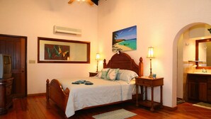 A bedroom with a wooden bed, a nightstand with a lamp, a painting on the wall, and a door. - Bayside Villa 3A (Roatan)
