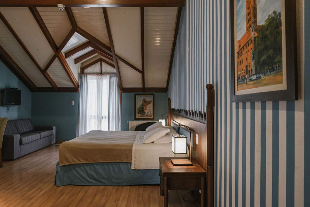 Un dormitorio con techo de madera, una cama con cabecera, una mesita de noche, una lámpara, un cuadro y una ventana con cortinas.