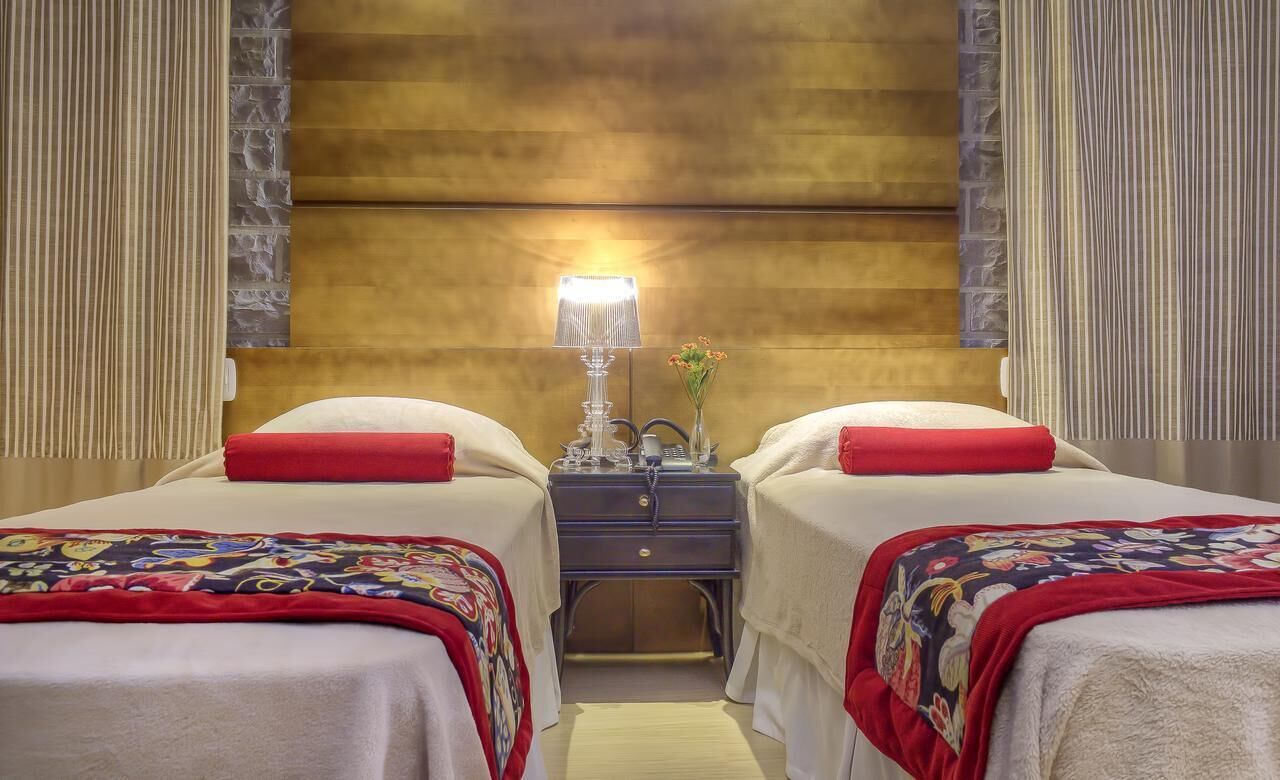 Habitación de hotel con dos camas, cada una con un cojín rojo y una manta colorida. Entre las camas, una mesita de noche con una lámpara y un jarrón de flores.