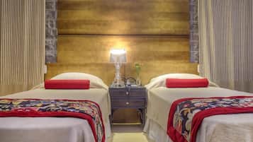 Quarto de hotel com duas camas, cada uma com almofada vermelha e manta colorida. Há uma mesa de cabeceira com abajur e um vaso de flores entre as camas.