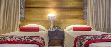 Habitación de hotel con dos camas, cada una con un cojín rojo y una manta colorida. Entre las camas, una mesita de noche con una lámpara y un jarrón de flores.