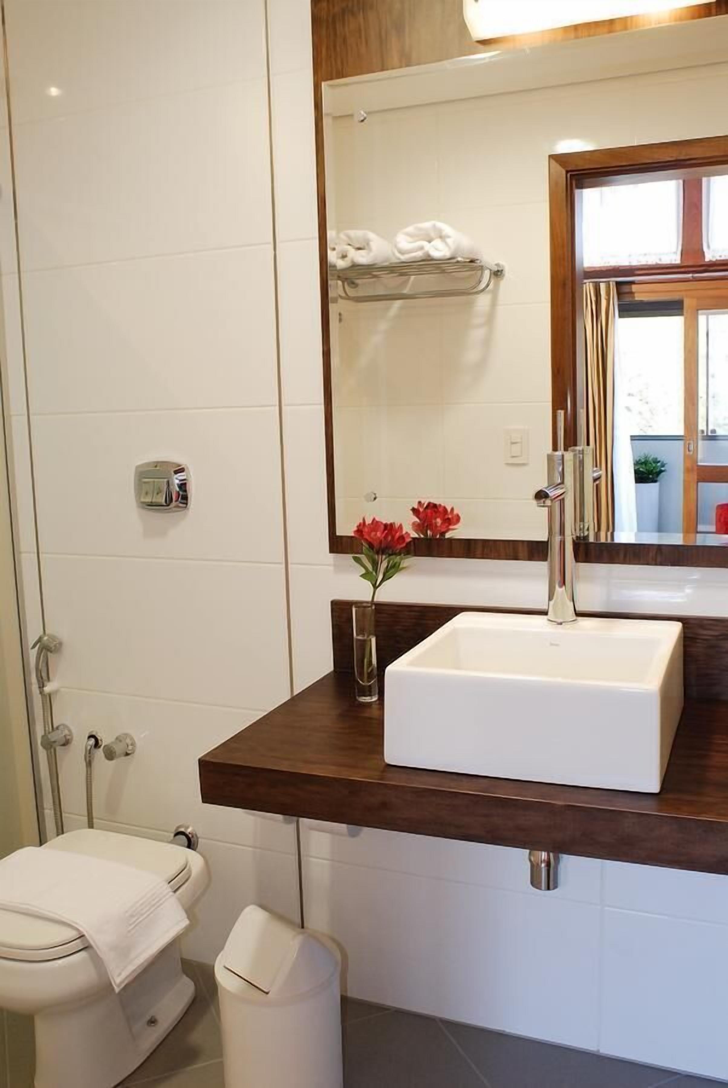 Un baño con inodoro blanco, un lavamanos blanco con encimera de madera, un espejo y un pequeño jarrón con flores rojas.
