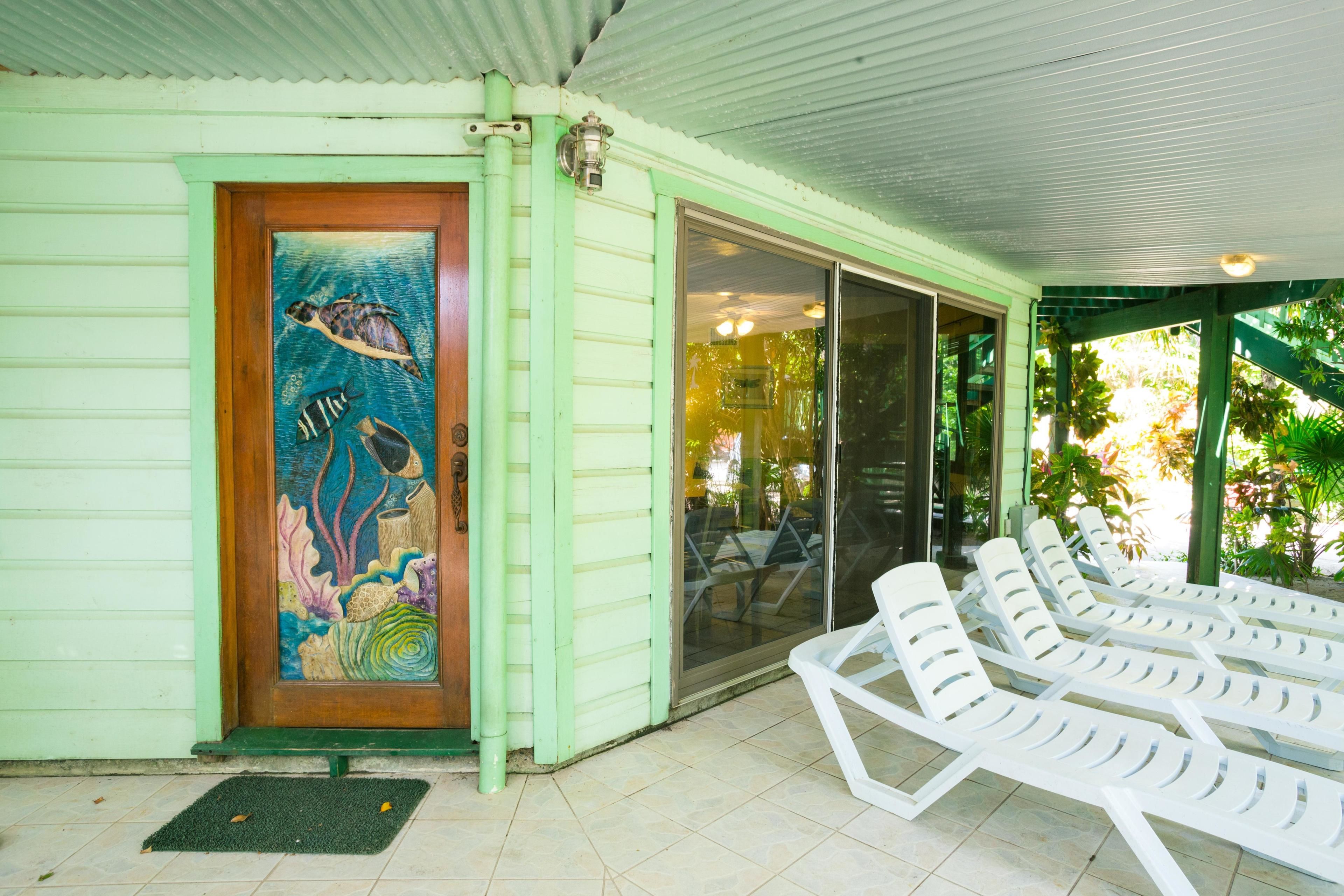 A covered outdoor area with a mural of a sea turtle on a door, white lounge chairs, and a glass door leading inside.