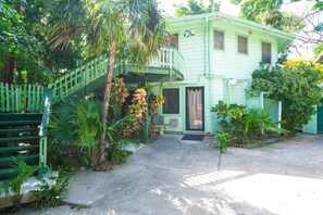 A light green house with a balcony, surrounded by lush greenery and a concrete driveway.