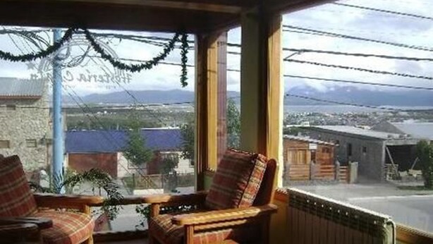 Una habitación acogedora con un banco y sillas de madera, una pequeña mesa con un periódico y vistas a un pueblo con edificios y montañas a lo lejos.