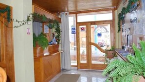 Zona de recepción con mostrador de madera, plantas en macetas y vista a la calle a través de puertas de vidrio.