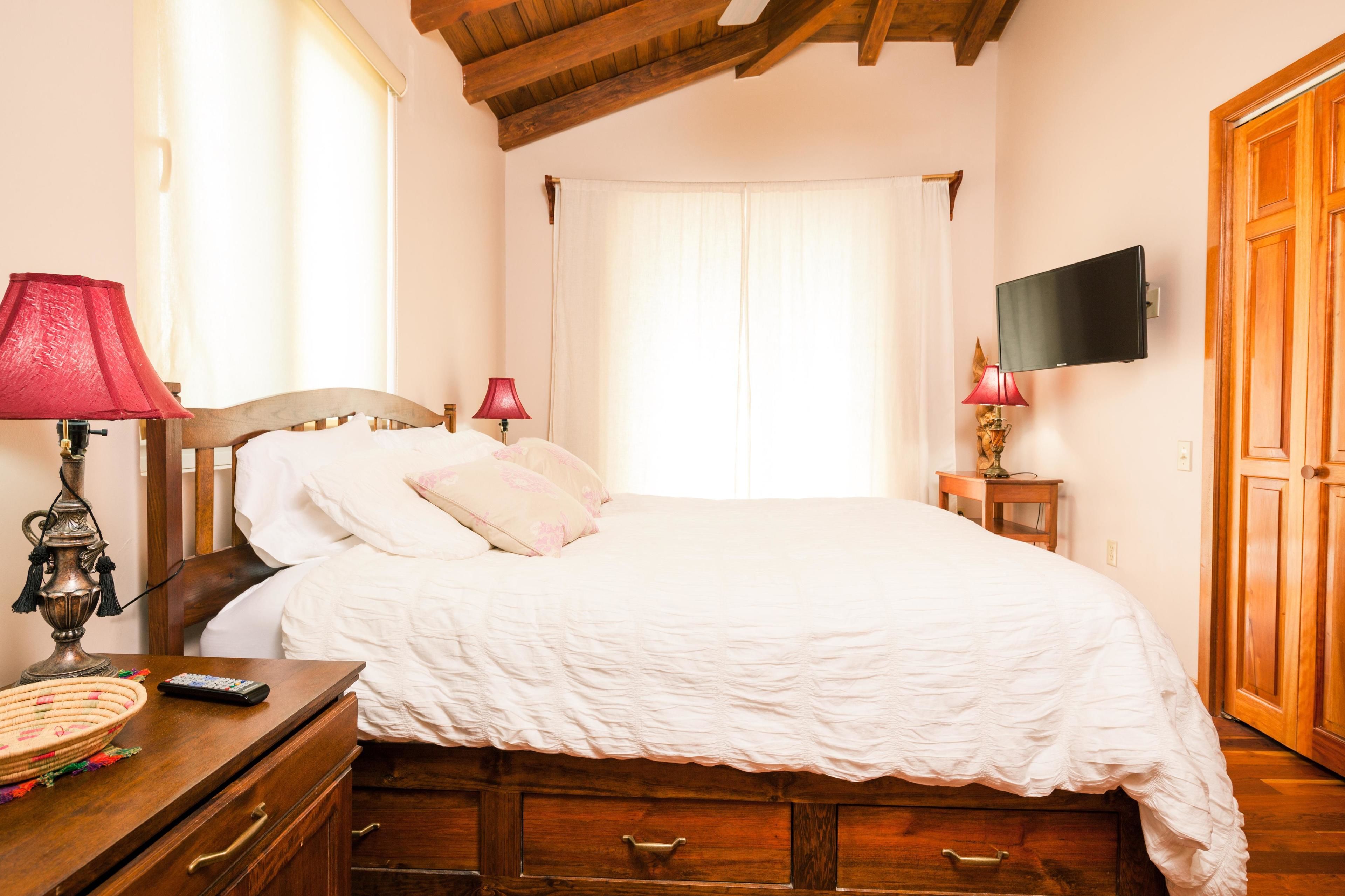 Une chambre à coucher avec un lit en bois, deux lampes de chevet, un téléviseur fixé au mur et une armoire en bois.