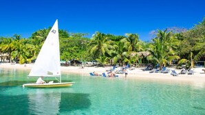 A sailboat with a white sail labeled "A3" on a clear turquoise sea, a sandy beach with palm trees, and sun loungers. - Coral Vista 1 2 bedroom option (Roatan)