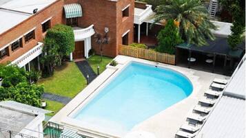 Ein rechteckiger Pool mit umlaufender Sonnenterrasse, ein Backsteinhaus mit überdachter Veranda und eine Palme.