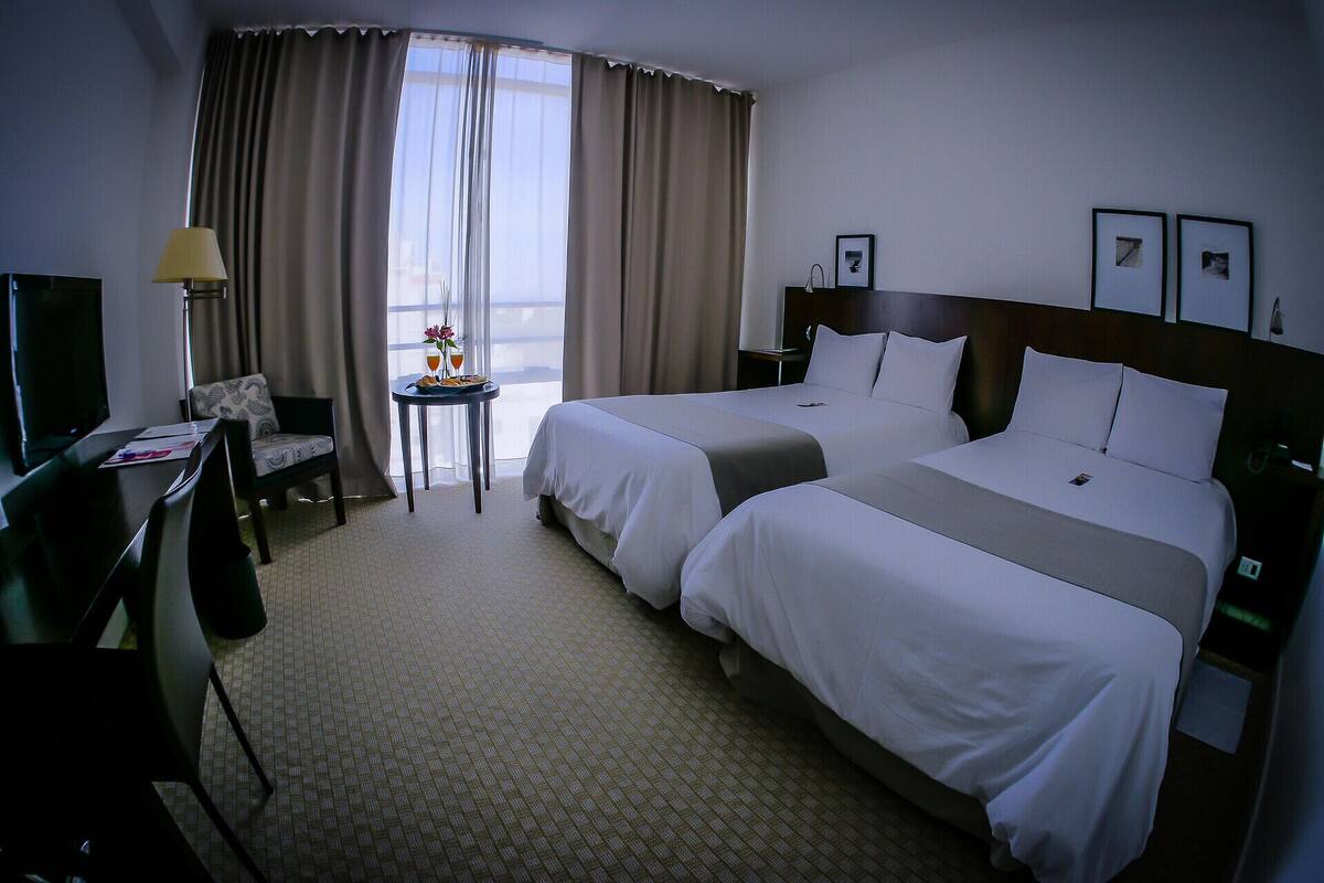 A hotel room with two beds, a desk, a chair, a table with drinks, and a TV.