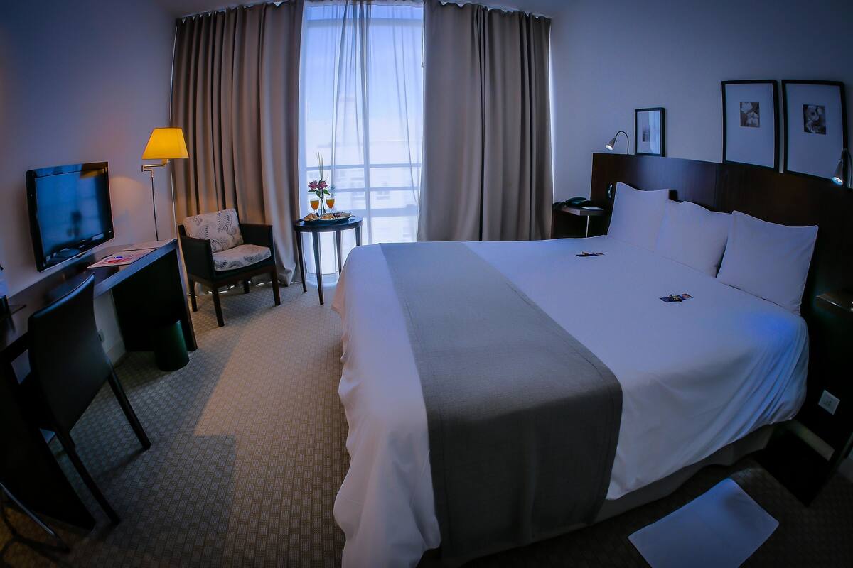 A hotel room with a large bed, a desk with a TV, a chair, a small table with drinks, and curtains.