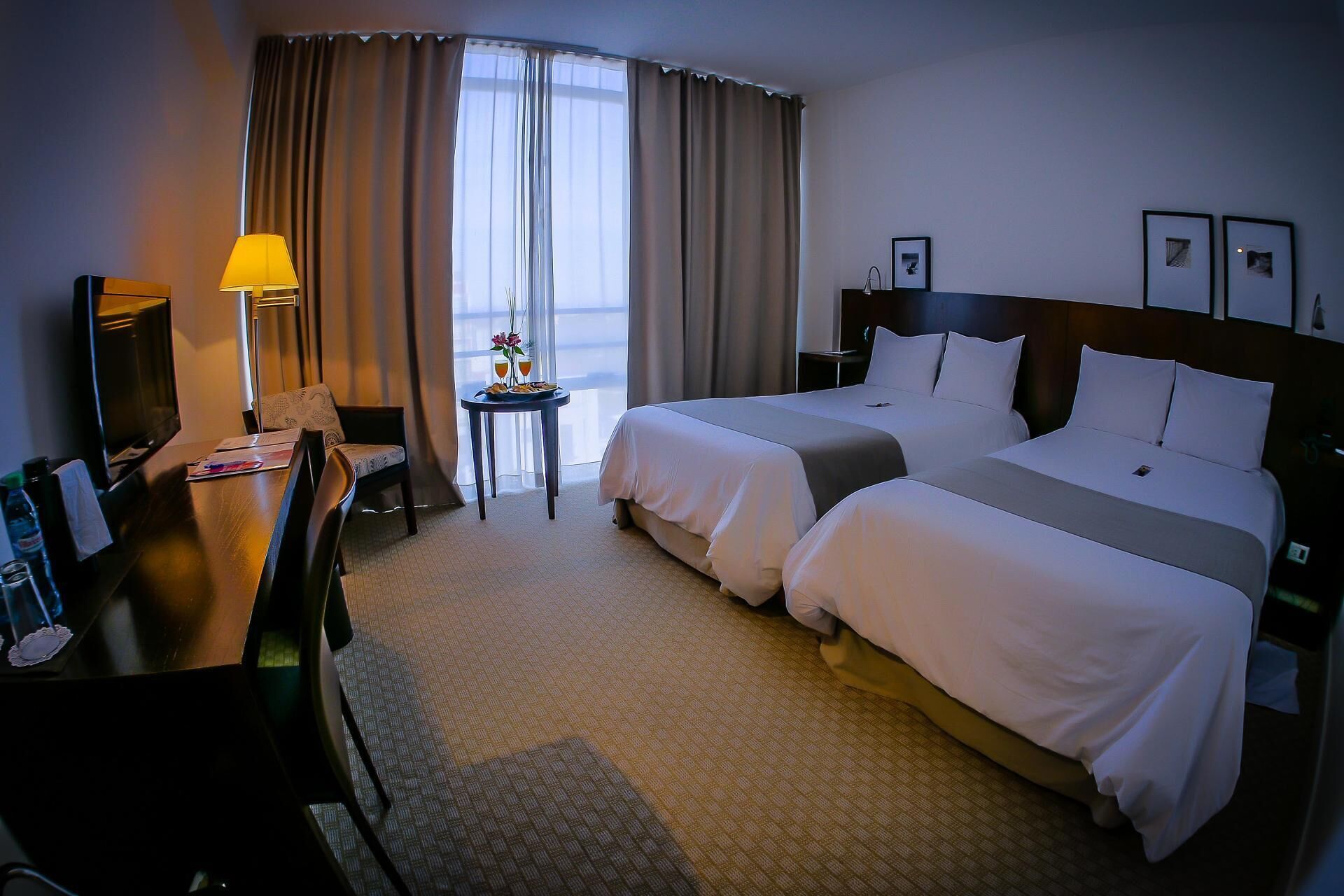 Quarto de hotel com duas camas, uma escrivaninha, uma cadeira, um abajur e uma pequena mesa com bebida e flores.