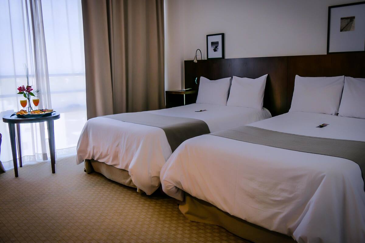 A hotel room with two beds, a small table with a drink and snacks, a nightstand, and a window with curtains.