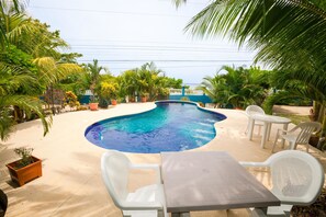 A swimming pool with a curved edge, surrounded by white plastic chairs and a table, with palm trees and potted plants around. - Sand Dollar (Roatan)