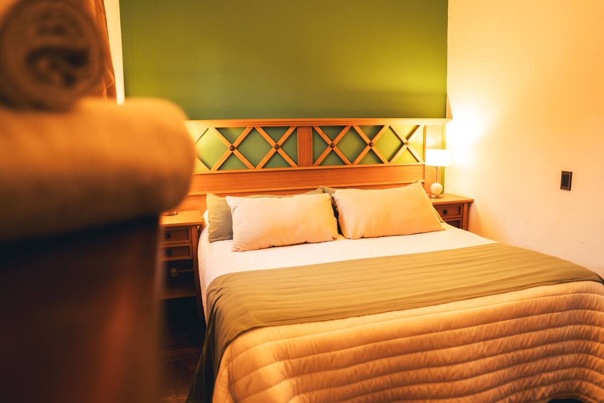 A hotel room with a neatly made bed, a wooden headboard with a geometric pattern, and a bedside table with a lamp.