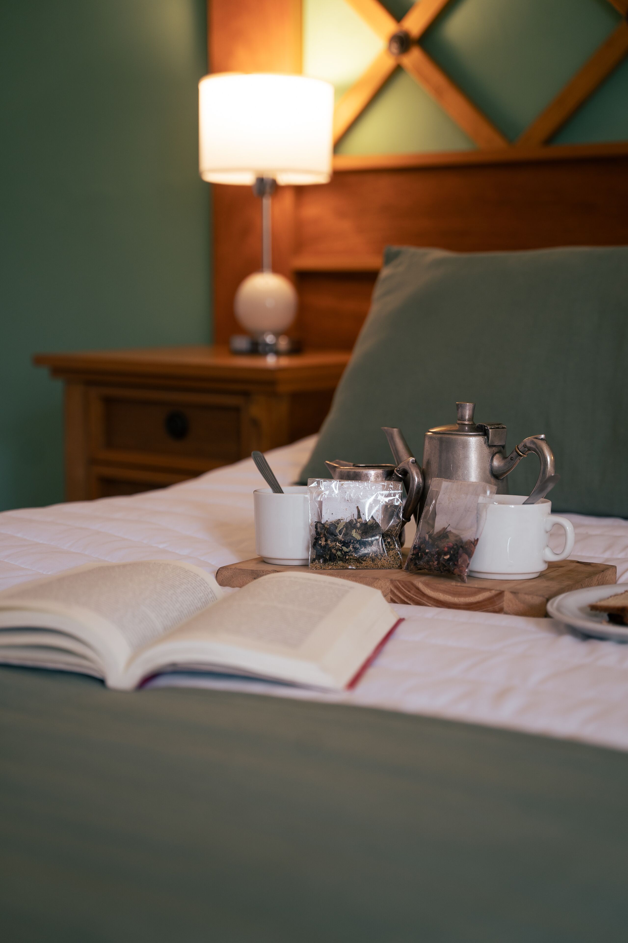 Una cama bien hecha con un libro, una tetera y dos tazas sobre una bandeja de madera.