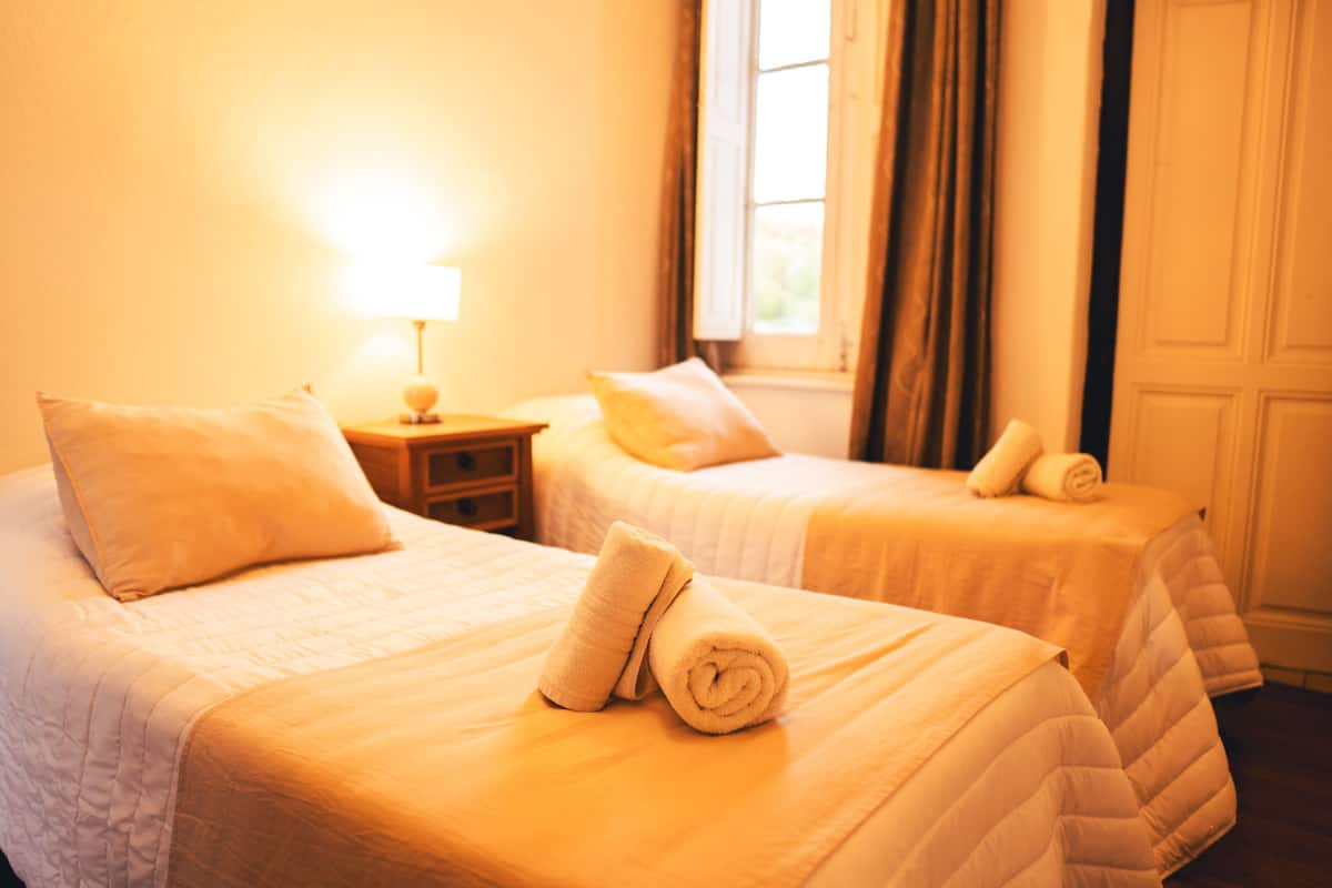Two single beds with white bedding and pillows, a nightstand with a lamp, and a window with curtains.