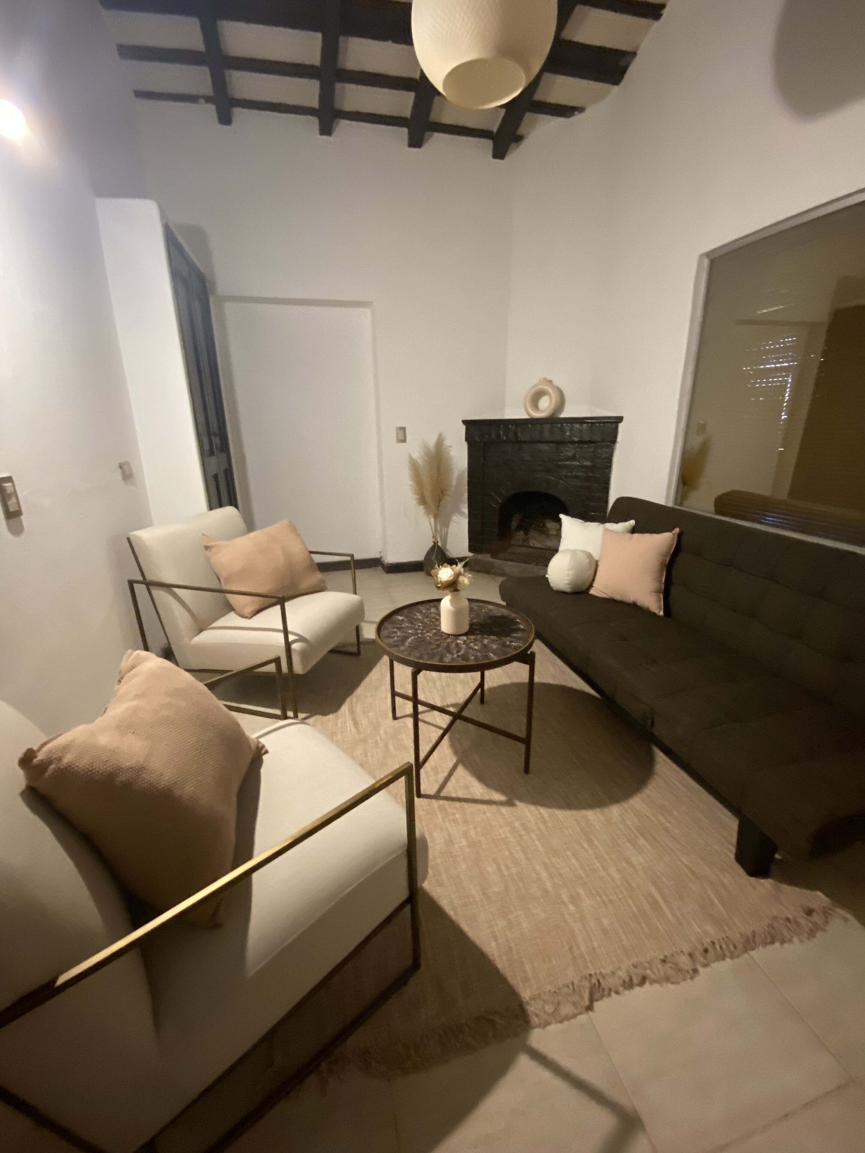 A modern living room with a dark-colored sofa, a white armchair, and a round coffee table. There is a fireplace with a decorative item on top.
