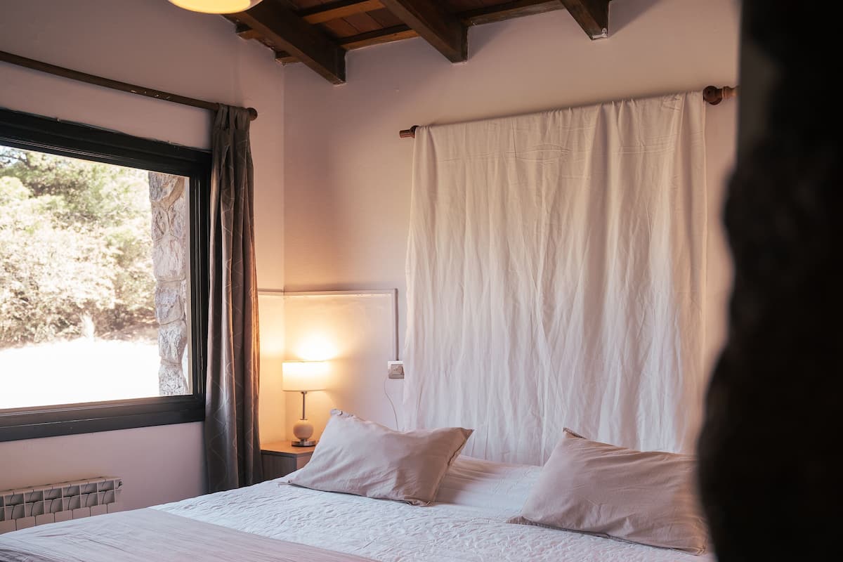 A bedroom with a large bed, a window with curtains, a lamp on a bedside table, and a wooden ceiling.