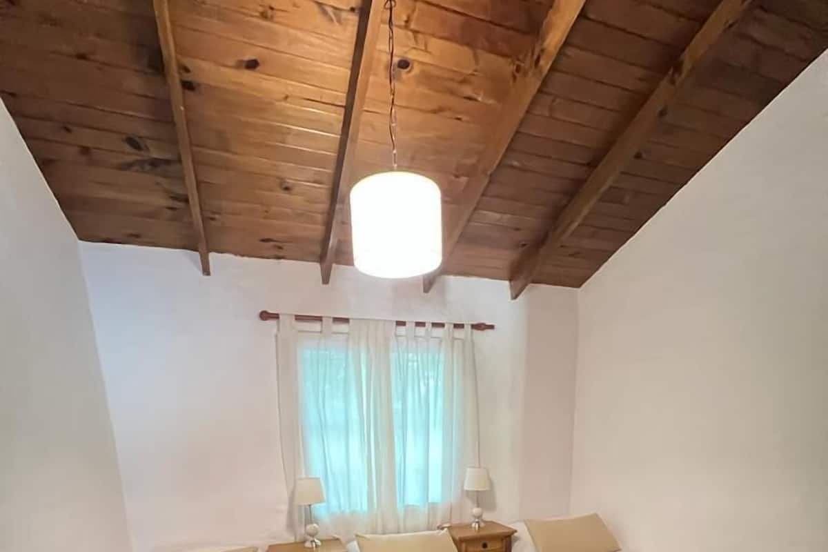 A small, cozy room with two single beds, a wooden ceiling, a window with curtains, and a hanging light fixture.
