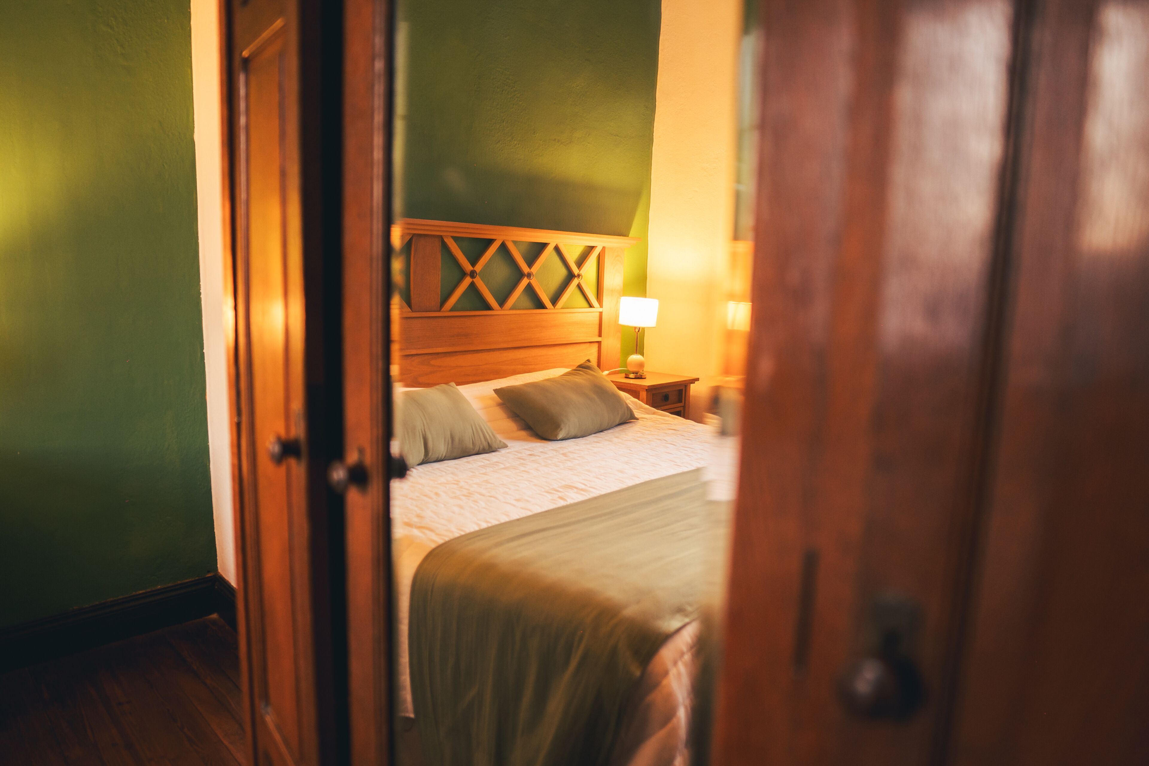 A hotel room with a wooden headboard, a bed with a green bedspread, and a nightstand with a lamp.