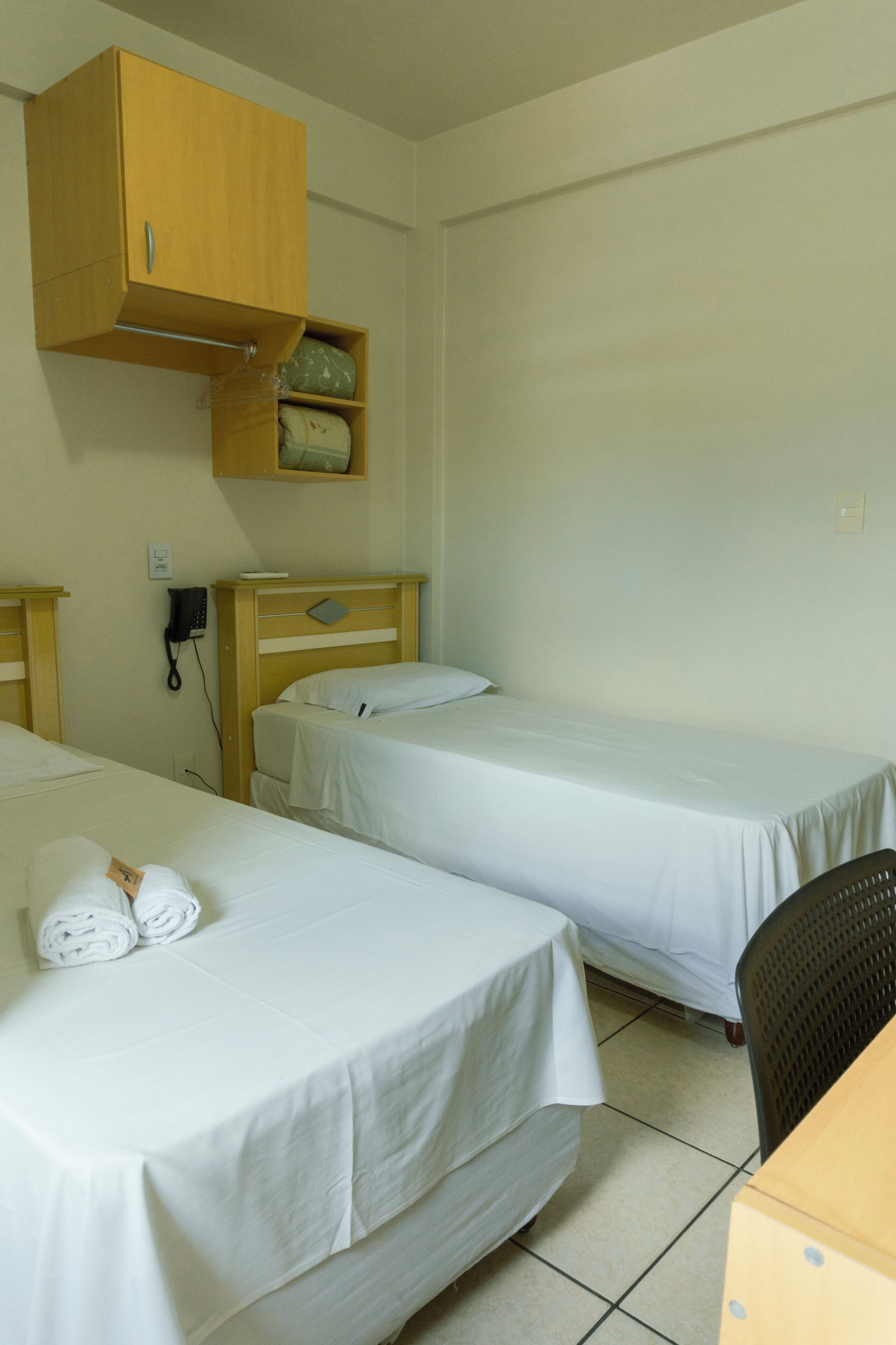 Una habitación pequeña y limpia con dos camas individuales, un armario de madera sobre la cama izquierda, un teléfono y una silla.