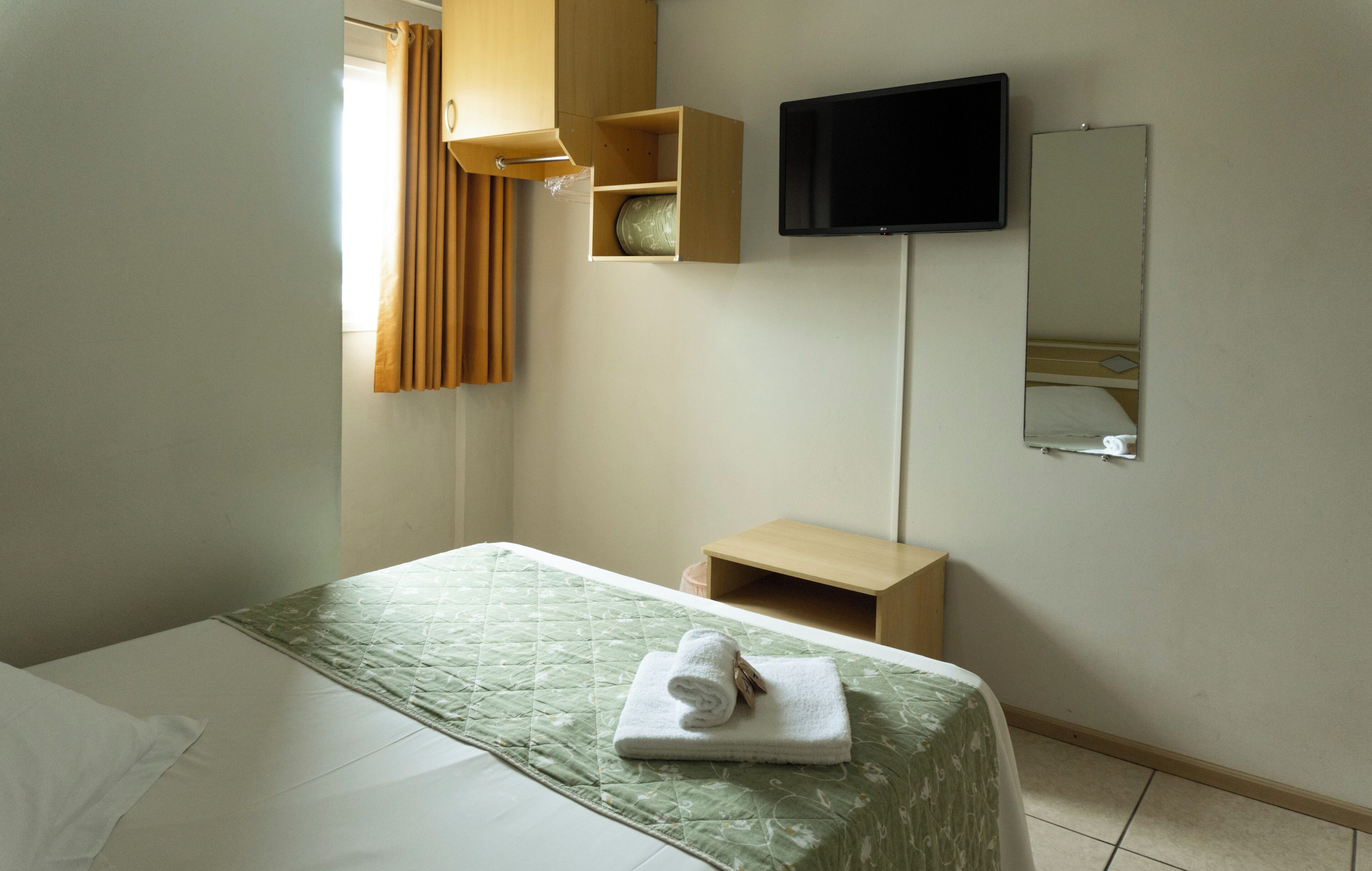 A hotel room with a bed, a television mounted on the wall, a mirror, and a small wooden shelf.