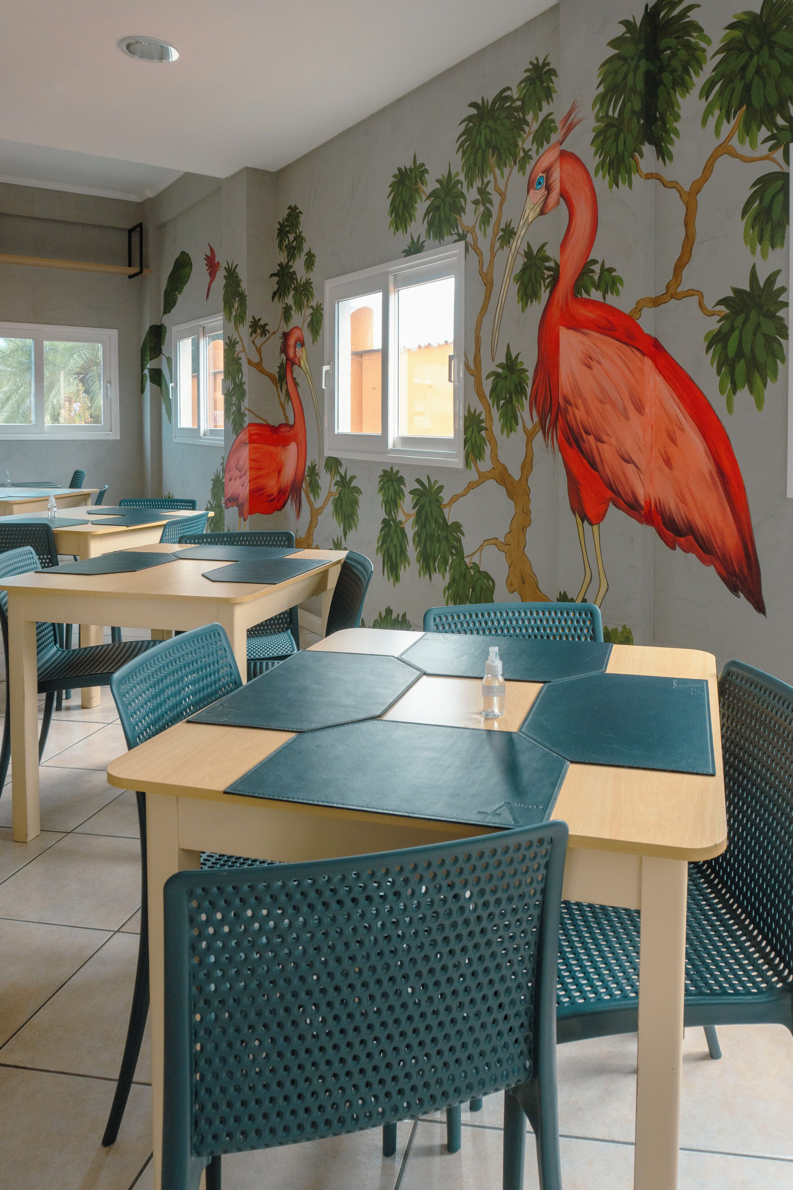 A dining area with tables and chairs, a mural of a red bird on the wall, and windows with natural light.