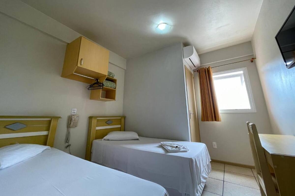 A small, clean room with two single beds, a wooden cabinet, a desk, and a window with curtains.