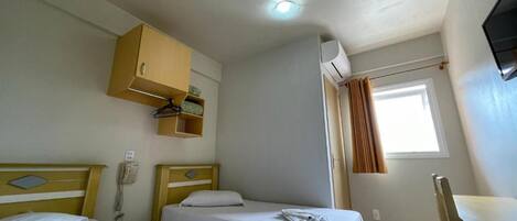 A small, clean room with two single beds, a wooden cabinet, a desk, and a window with curtains.