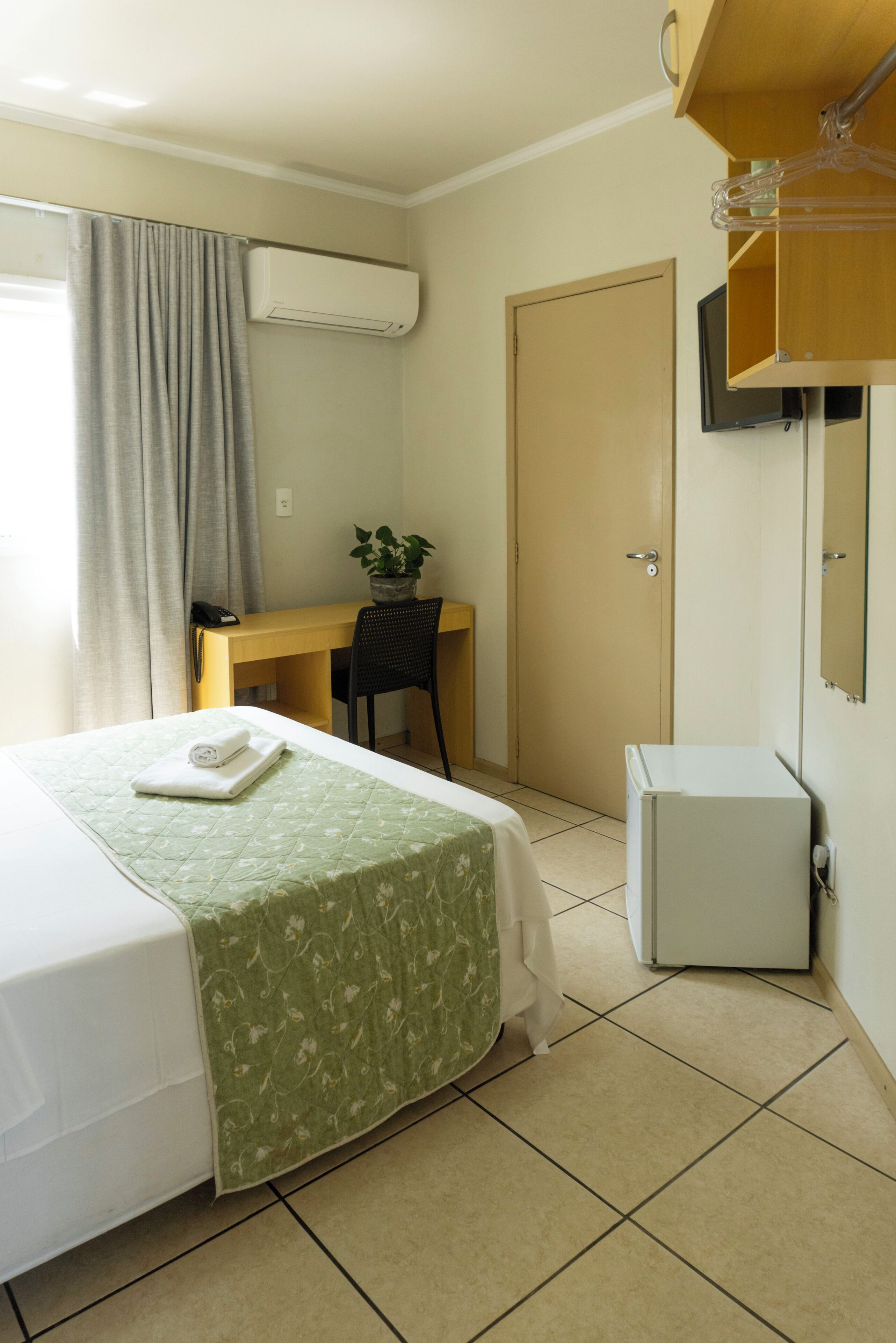 A hotel room with a bed, a desk, a chair, a small cabinet, and an air conditioning unit.