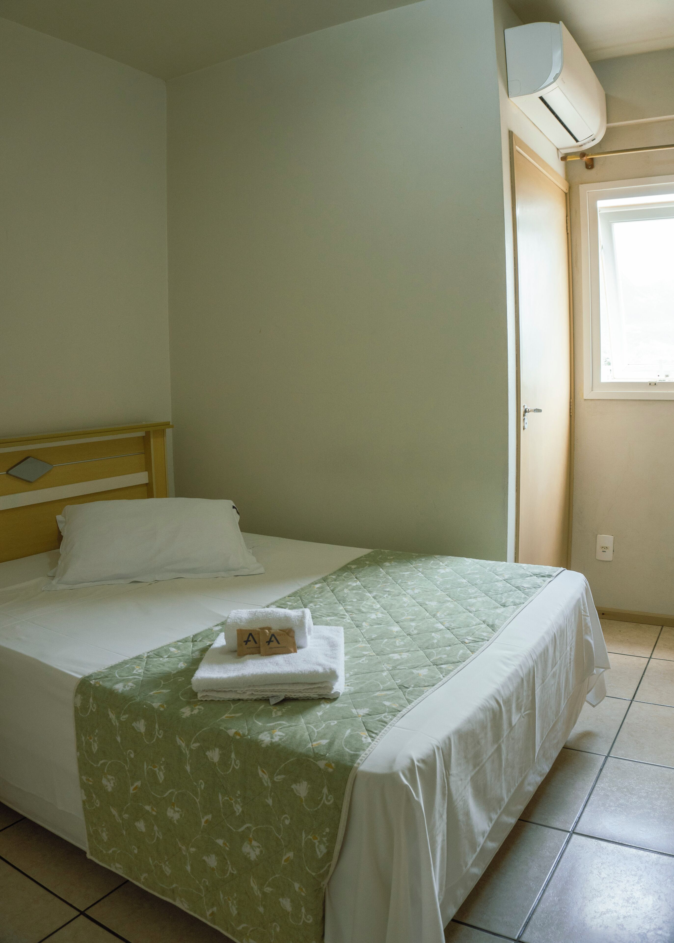 Una cama bien hecha con una colcha floral verde y ropa de cama blanca, un cabecero de madera y un equipo de aire acondicionado montado en la pared.