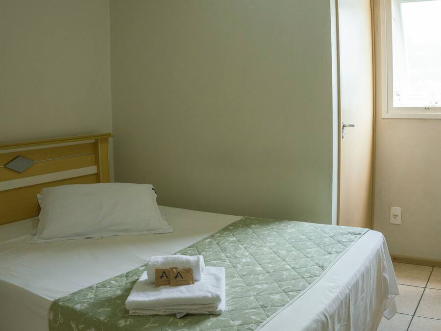 A neatly made bed with a green floral bedspread and white linens, a wooden headboard, and an air conditioning unit mounted on the wall.