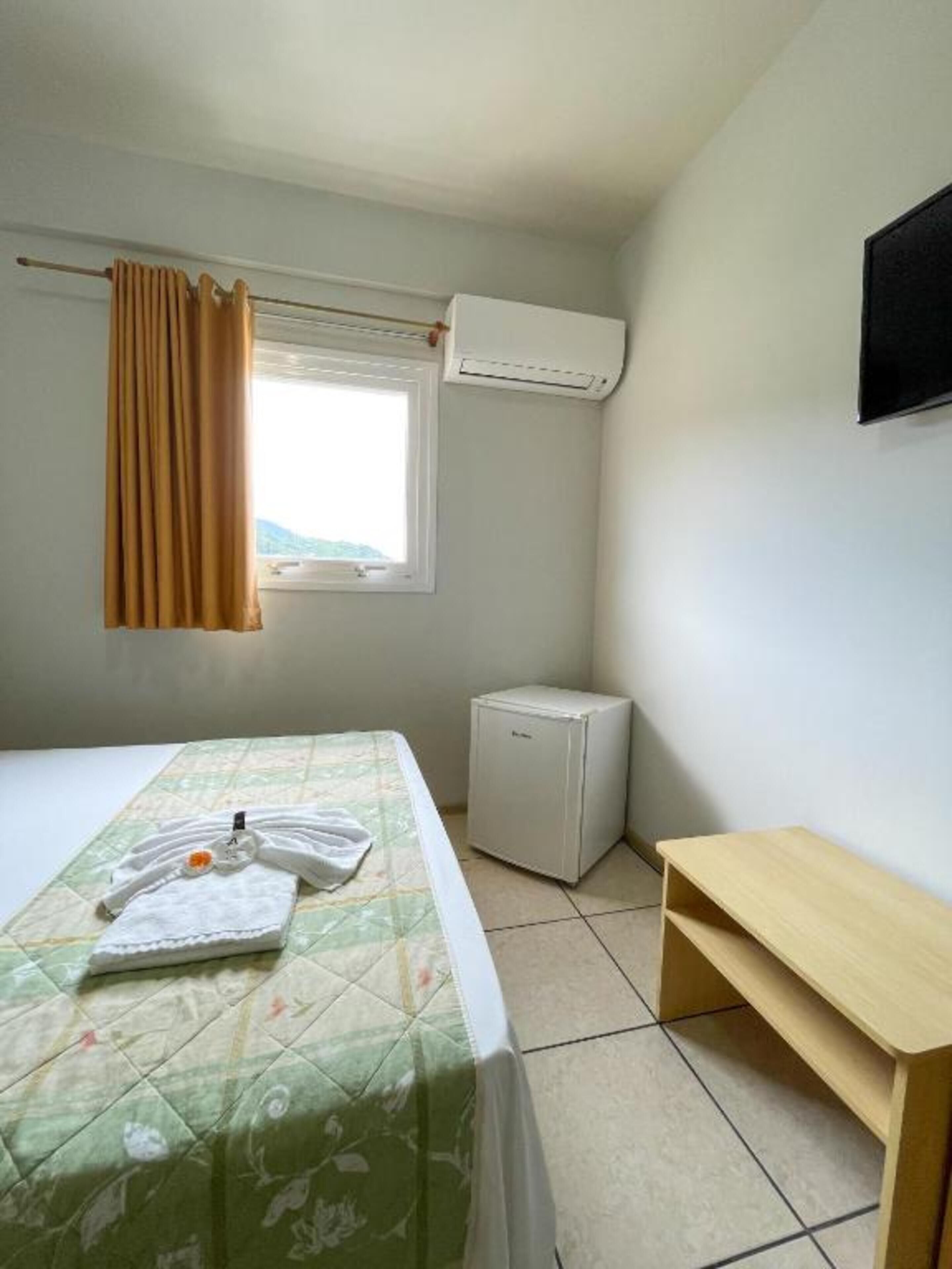 A small, simple room with a bed, a small table, an air conditioning unit, and a window with curtains.