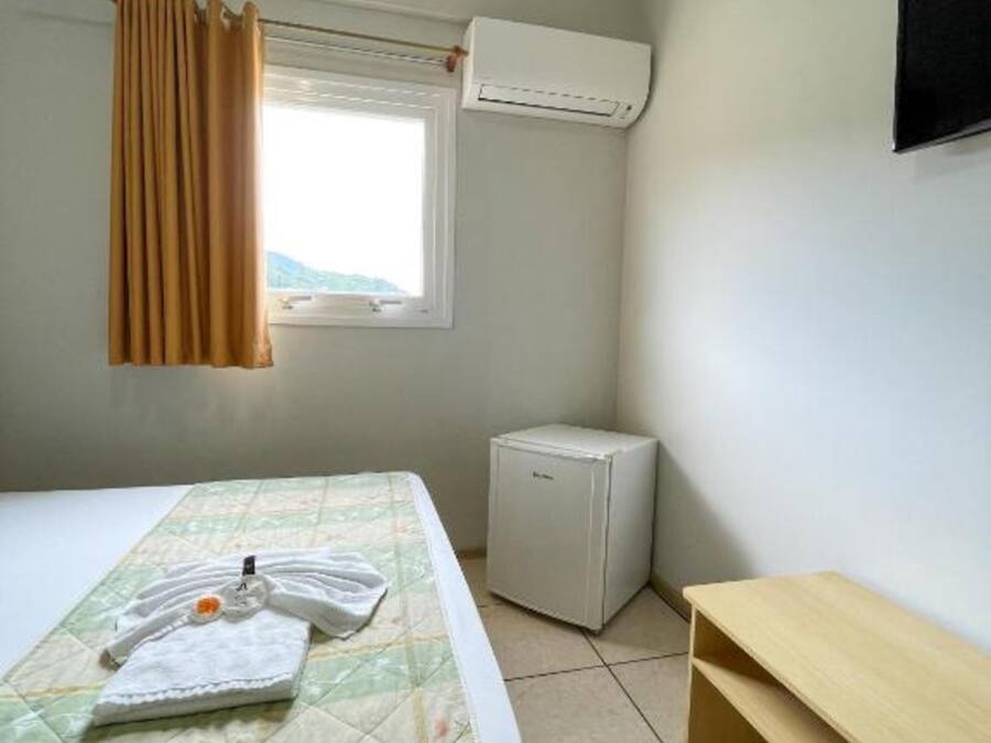A small, simple room with a bed, a small table, an air conditioning unit, and a window with curtains.