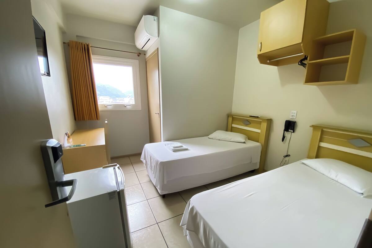 A hotel room with two beds, a small desk, a refrigerator, a window with curtains, and a wall-mounted phone.