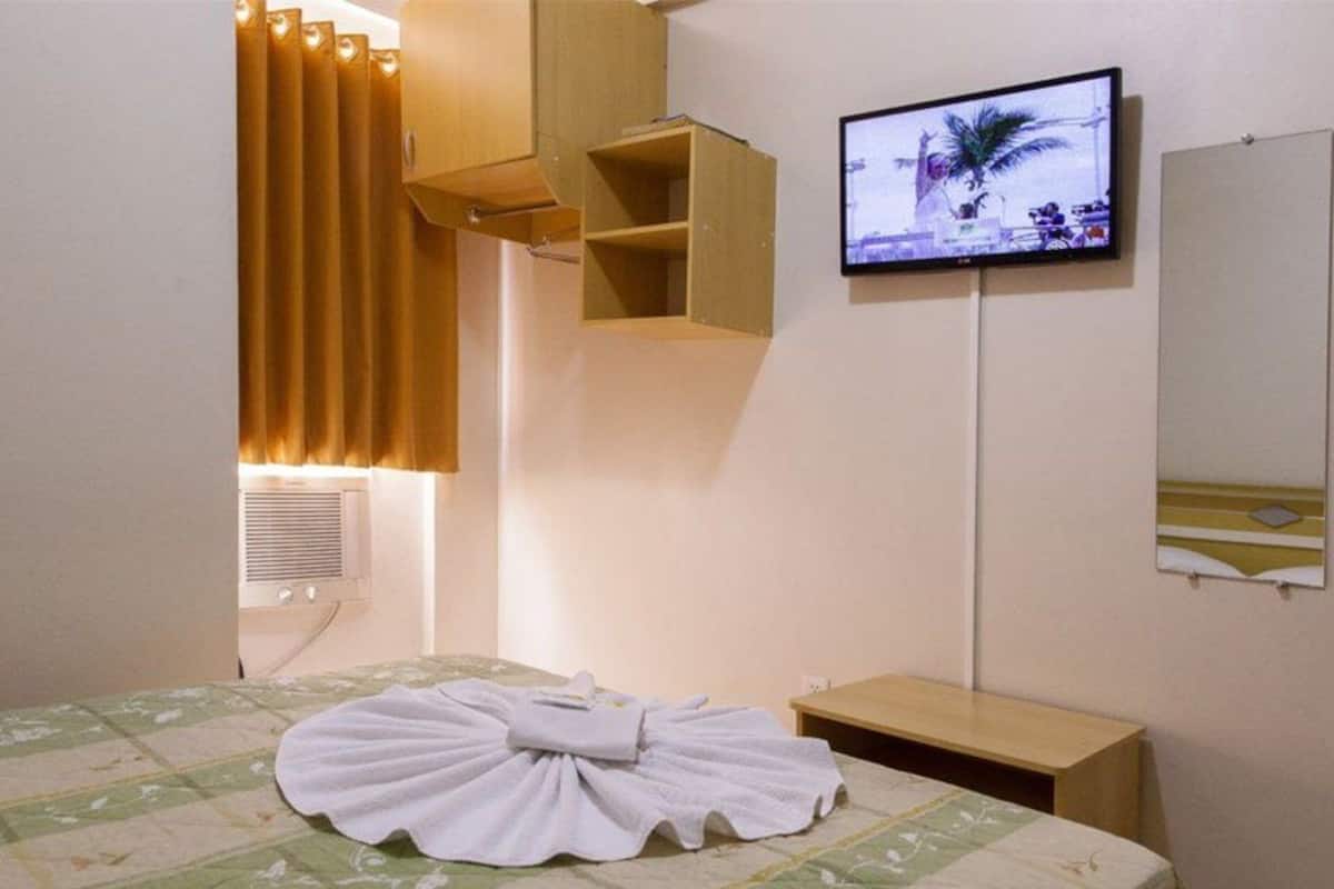 A hotel room with a bed, a TV mounted on the wall, a mirror, and a window with curtains.