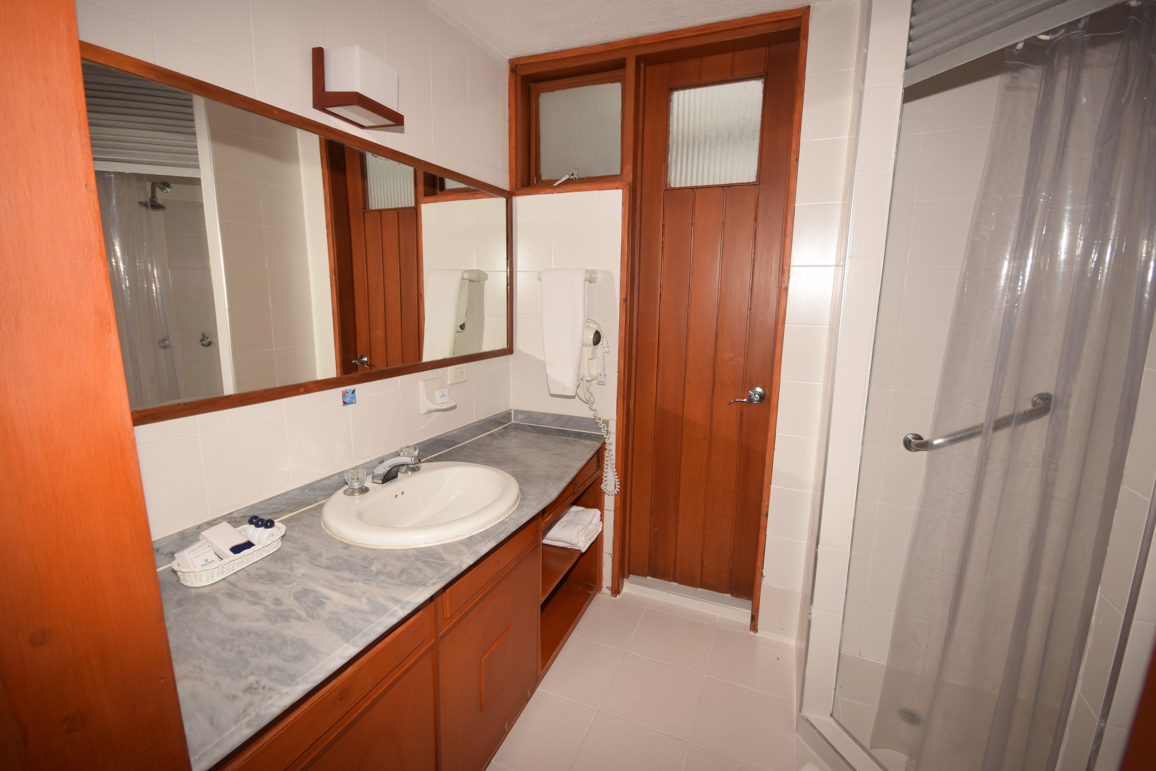 A bathroom with a marble countertop, a sink, a mirror, wooden cabinets, a shower with a glass door, and a towel rack.
