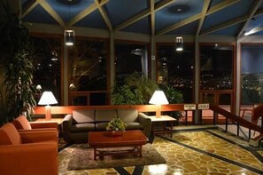 Ein großzügiger Aufenthaltsraum mit hoher Decke, großen Fenstern mit Stadtblick und einer Sitzecke inklusive Sofa und Sesseln.