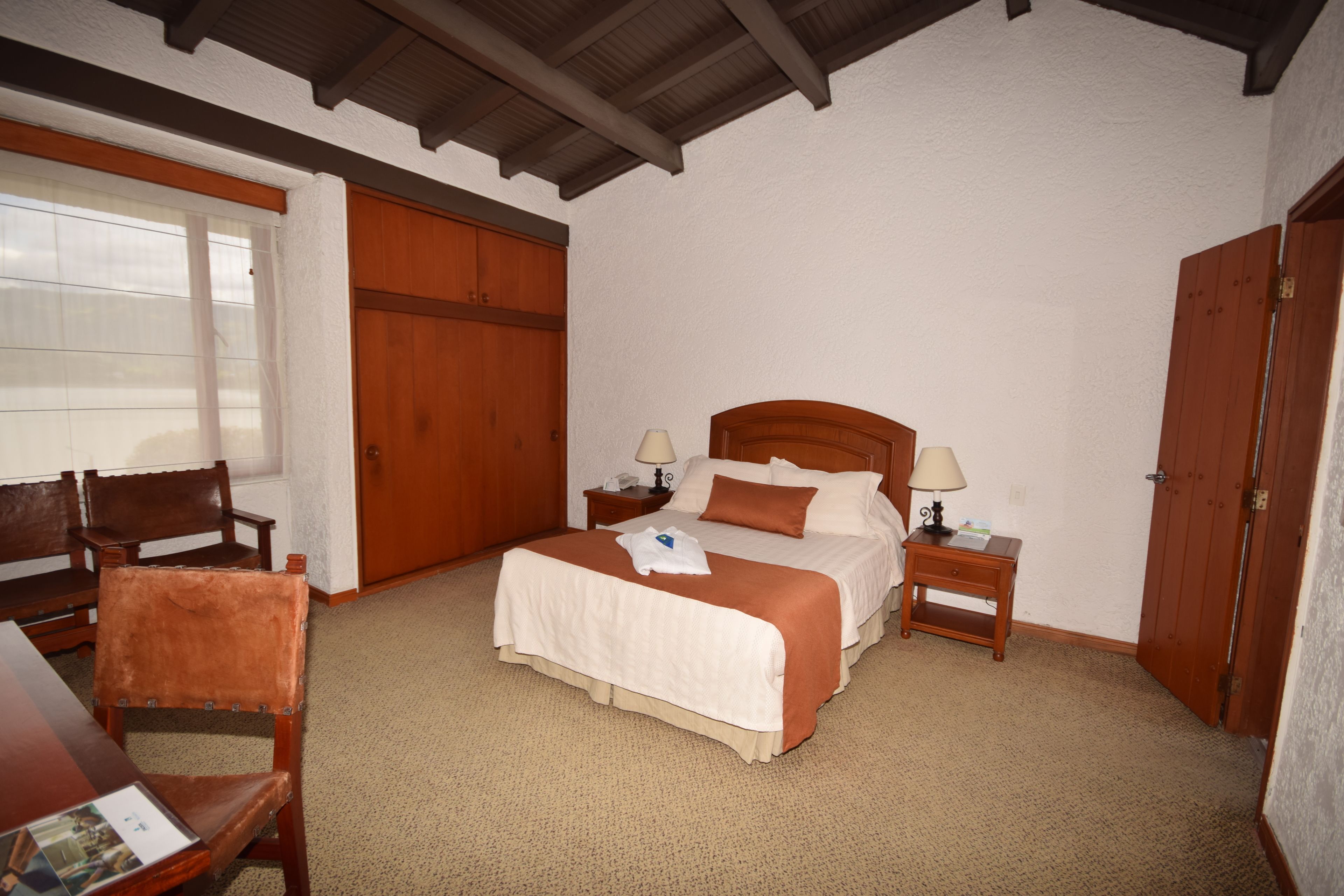 A hotel room with a wooden bed, two bedside tables with lamps, a wooden chair, and a large window with blinds.