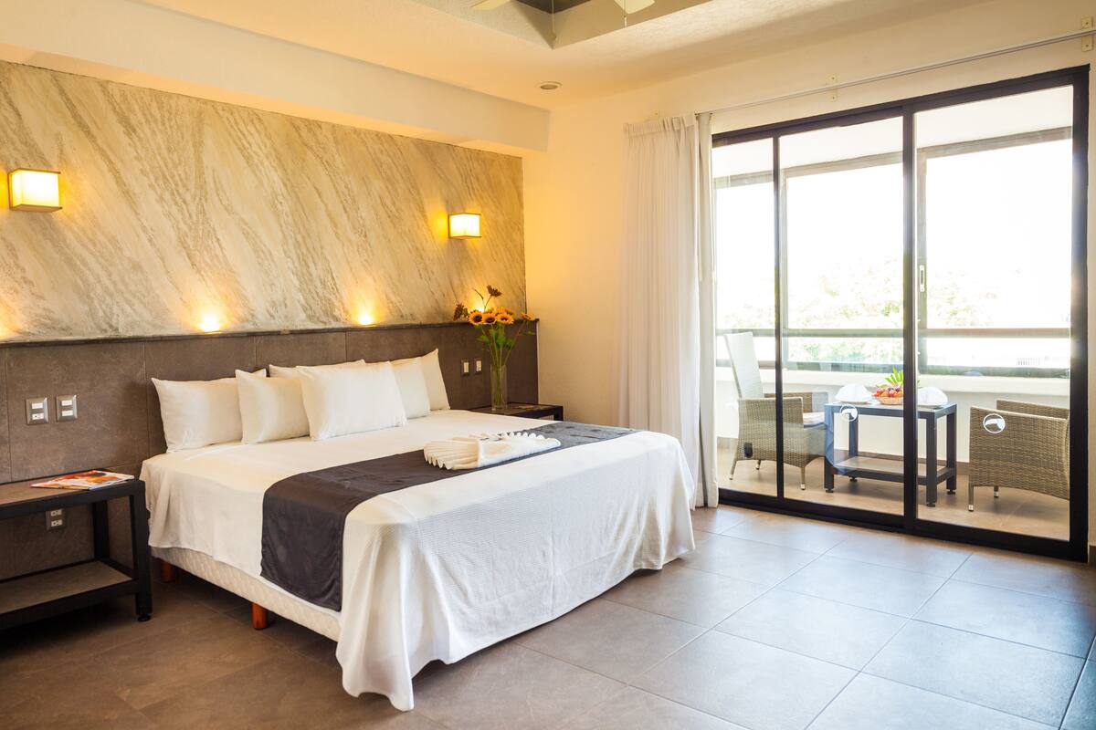 Habitación de hotel con una cama grande, una mesita de noche, una mesa con sillas y vistas a través de un ventanal corredero.