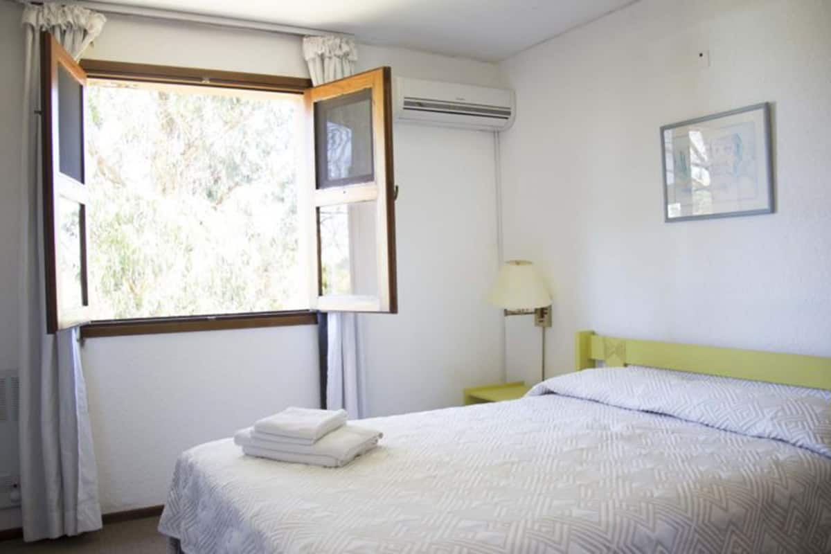 A hotel room with a bed, a nightstand, a lamp, an air conditioning unit, and a window with curtains.