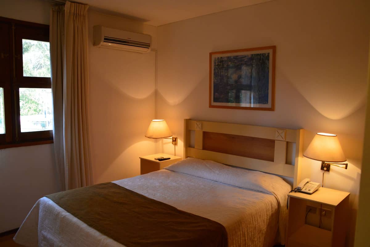 A hotel room with a bed, two bedside lamps, a painting on the wall, and an air conditioning unit.