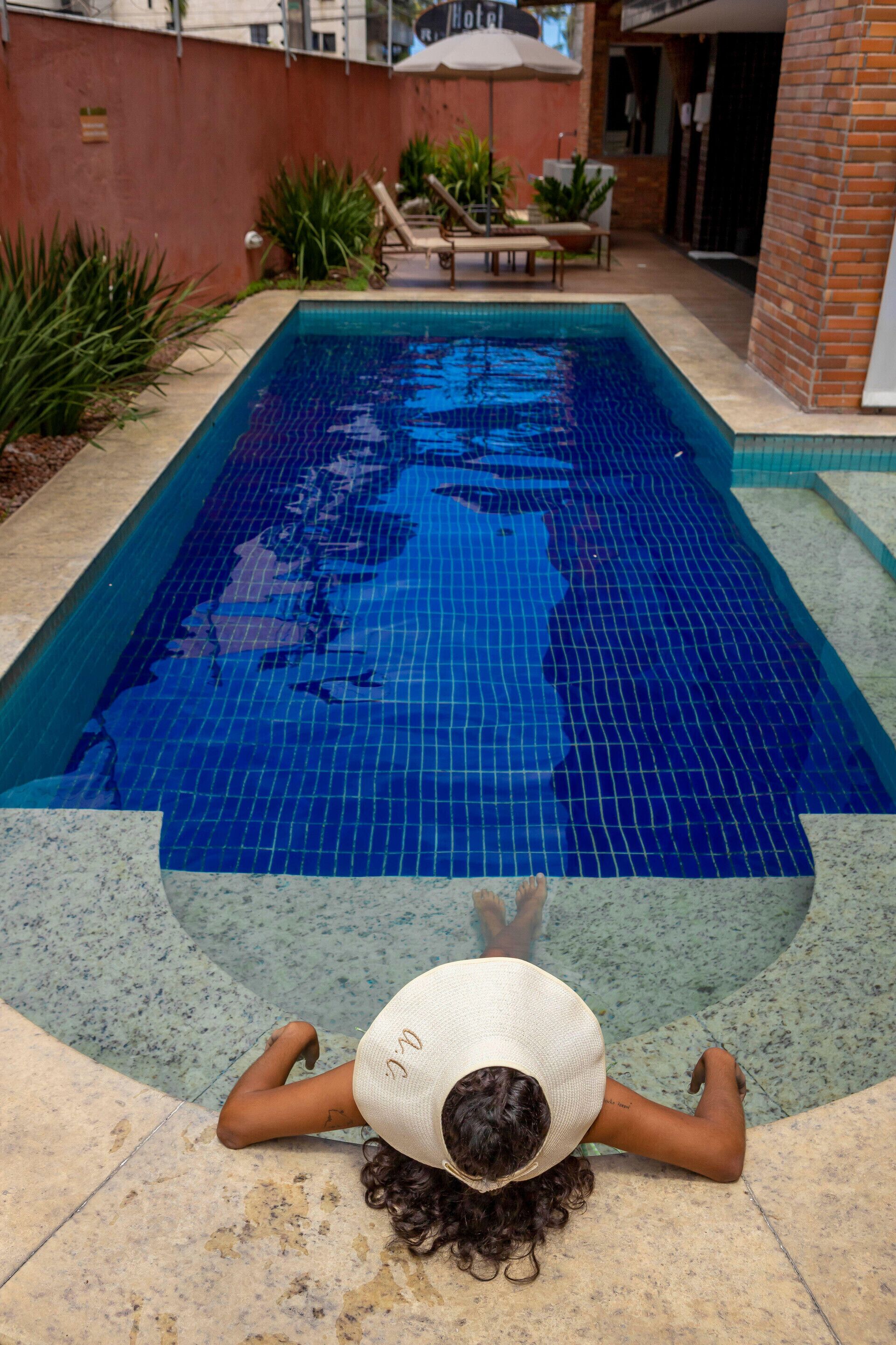 Una piscina con borde de baldosas, rodeada de plantas y un muro de ladrillo.